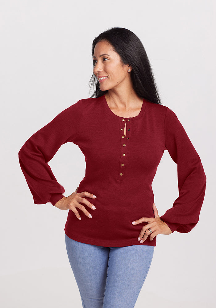 A woman with long dark hair smiles and looks to her left. She's wearing a Sweet Pepper Charlotte Puff Sleeve Top from Woolx and light blue jeans, with her right hand resting on her hip against the plain gray background.