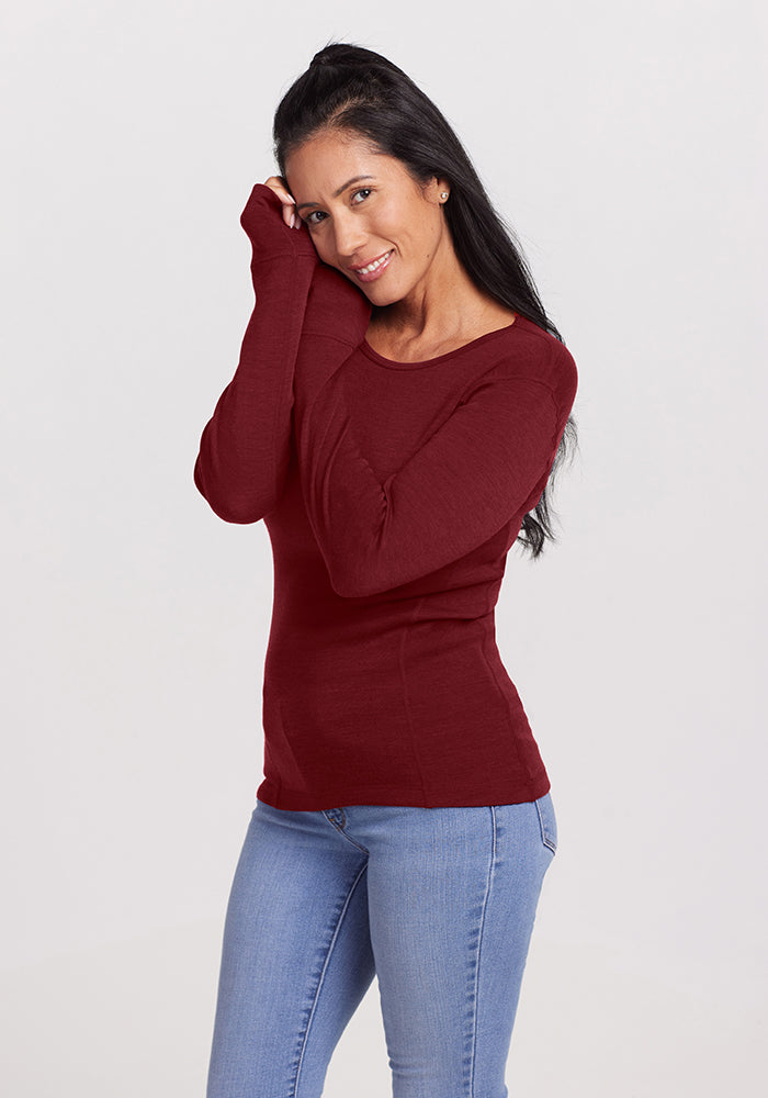 A woman with long, dark hair smiles while wearing a Sweet Pepper Hannah Long Sleeve Top by Woolx and light blue jeans. She stands casually against a plain, light background, her right hand gently resting on her cheek.