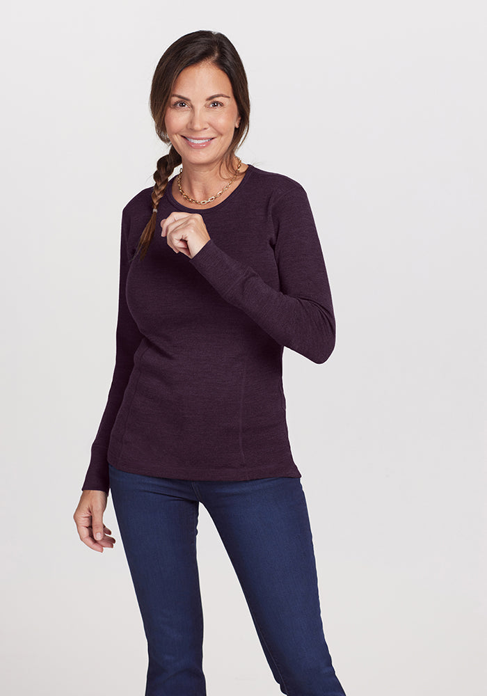 A woman with long dark braided hair smiles against a plain background, wearing the Woolx Hannah Long Sleeve Top in Deep Plum and blue jeans, with a necklace. She holds a lock of her hair in her right hand.