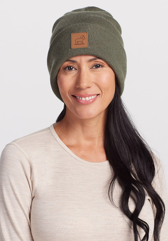 A person with long, dark hair is smiling and wearing a Woolx Baylor Beanie in Spruce Heather, which features a small patch on the front. The individual is also dressed in a light-colored, long-sleeve shirt. The background is plain white.