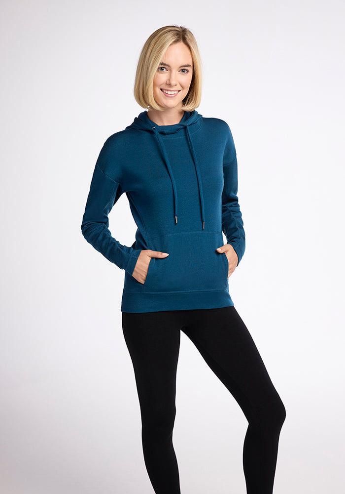 A woman with shoulder-length blond hair is standing and posing in a relaxed manner. She is wearing the Woolx Callie Hoodie in Real Teal, featuring a front pocket, and black leggings, embodying slouchy street style. She is smiling and holding her hands in the hoodie pocket against a plain white background.