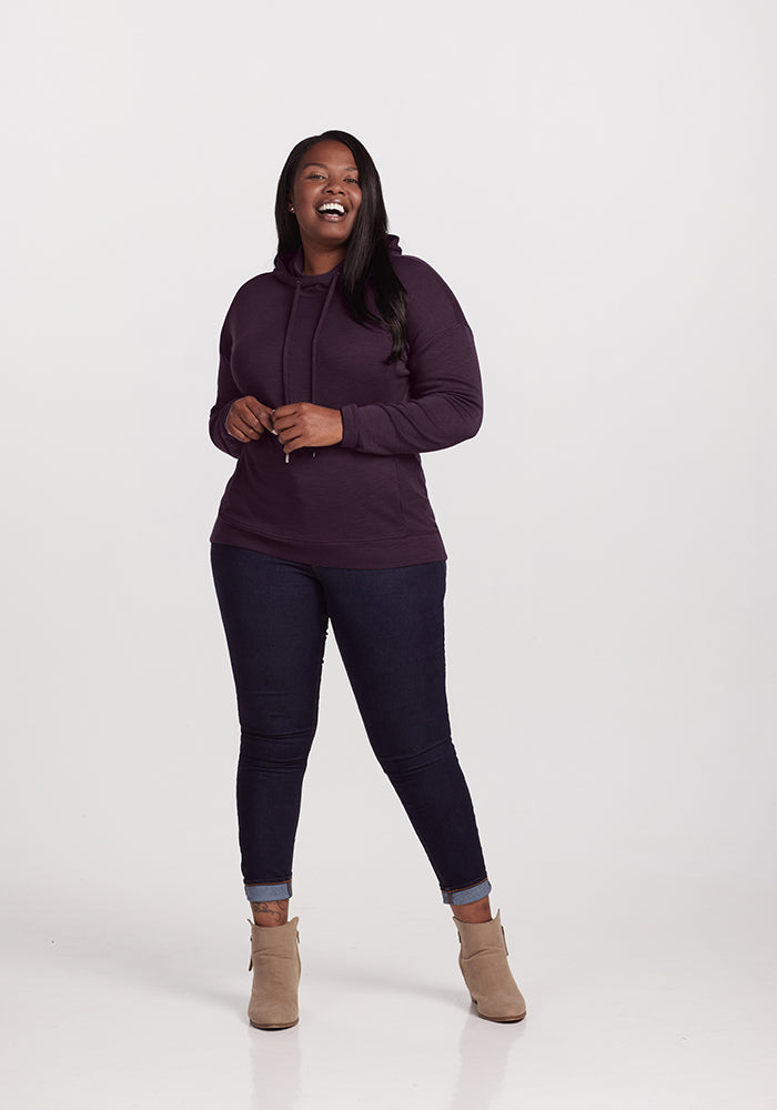 A woman stands smiling against a white background, showcasing her street style. She is wearing the Callie Hoodie in Deep Plum from Woolx, made from luxurious Australian Merino Wool. Paired with dark blue jeans with rolled cuffs and beige ankle boots, her long dark hair flows over her shoulders as she casually rests her hands in front of her.