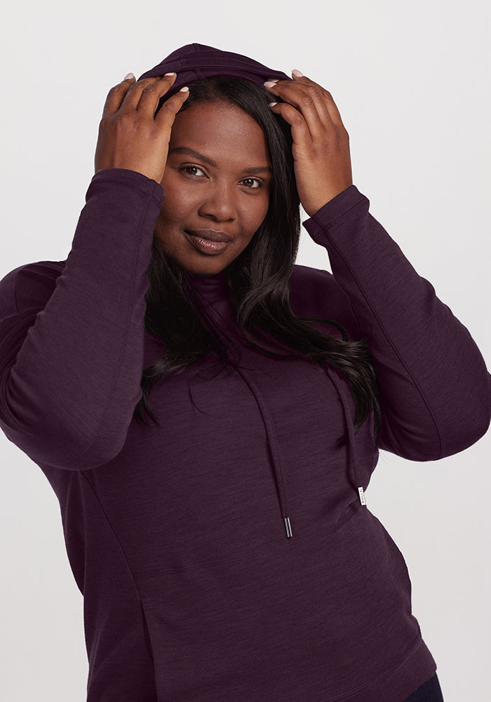 A person with long dark hair wears a Woolx Callie Hoodie in Deep Plum, adjusting the hood with both hands. They are looking directly at the camera with a slight smile. The street style look is effortlessly chic against the plain, light-colored background.