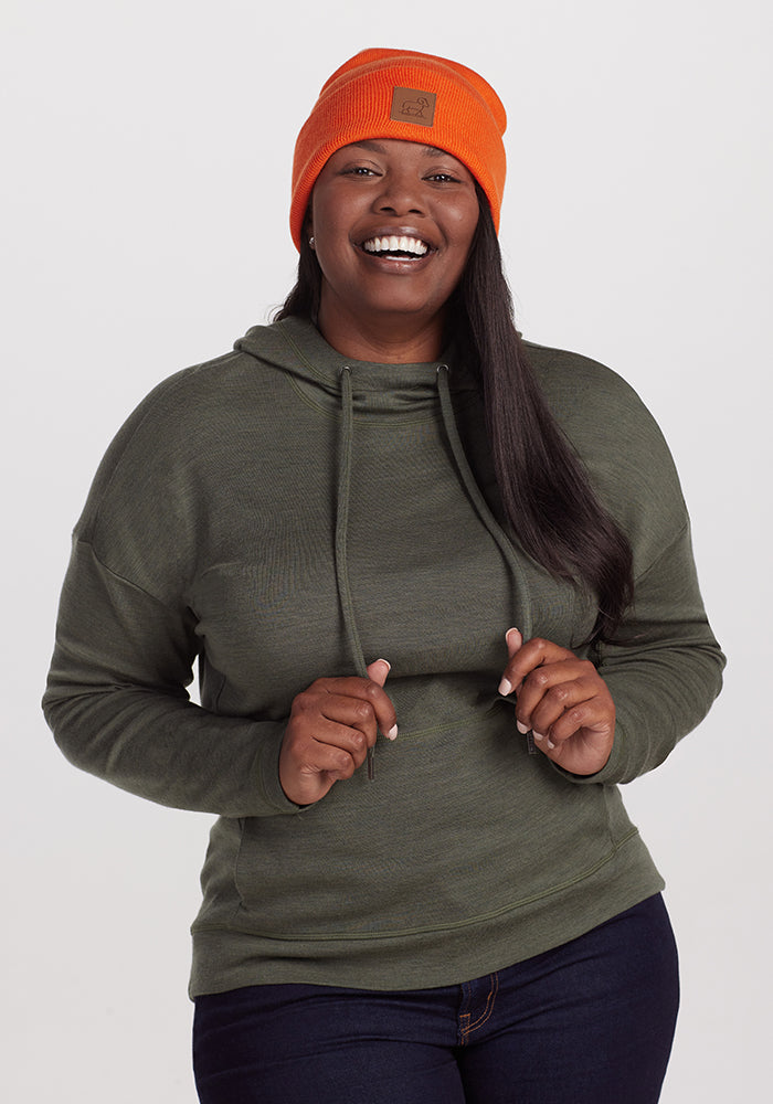 A person wearing a dark green hoodie and a Woolx Baylor Beanie in bright orange is standing against a plain background. They have long, dark hair and are smiling while holding the hoodie strings with both hands.