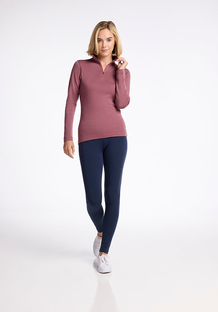 A woman with shoulder-length blonde hair is walking towards the camera. She is sporting the Woolx Brooke 1/4 Zip in maroon, paired with navy blue leggings and white sneakers. With a slight smile, she touches the zipper of her top with her left hand.