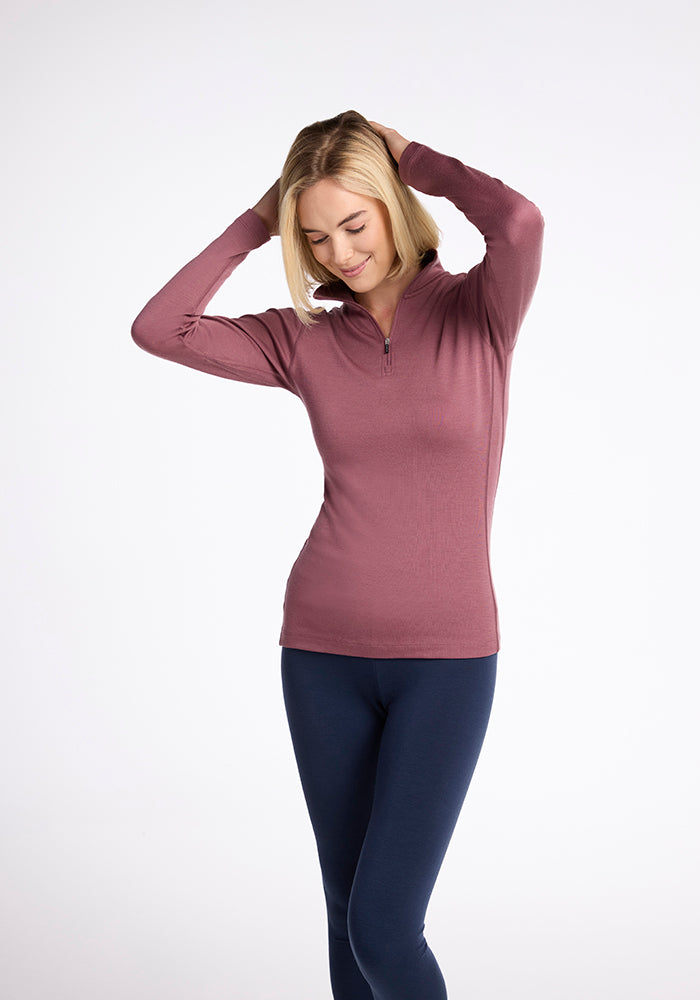 A woman with blonde hair stands against a plain white background, wearing a Woolx Brooke 1/4 Zip top in mauve and navy blue leggings. She has her hands raised near her head and is looking down with a slight smile.