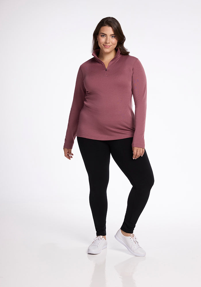 A person is standing against a plain white background, wearing the Brooke 1/4 Zip from Woolx in a pinkish-purple hue, paired with black leggings and white sneakers. The individual has medium-length hair and is smiling slightly while looking at the camera.