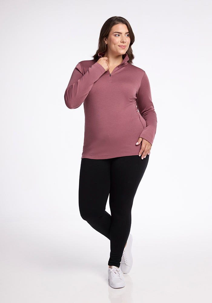 A confident person poses in a Woolx Brooke 1/4 Zip long-sleeve top in mauve and black leggings. They are slightly smiling and touching their chin with one hand, while their other hand rests on their hip. They wear white sneakers and stand against a plain white background.