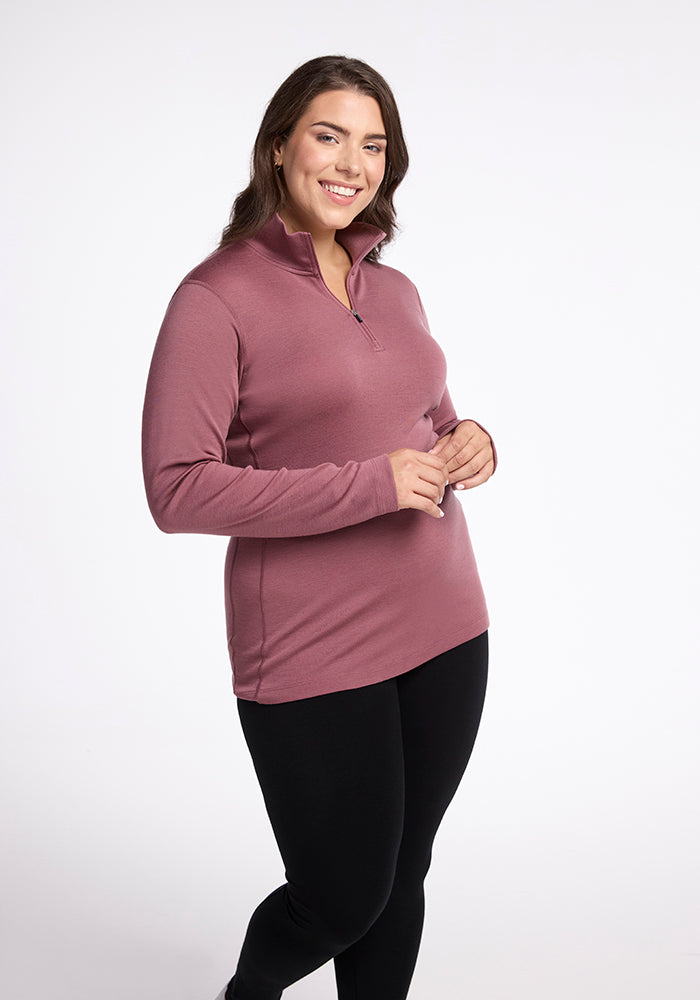 A woman with long brown hair smiles while standing. She is wearing the Brooke 1/4 Zip in dusty rose from Woolx and black leggings. The background is plain white.