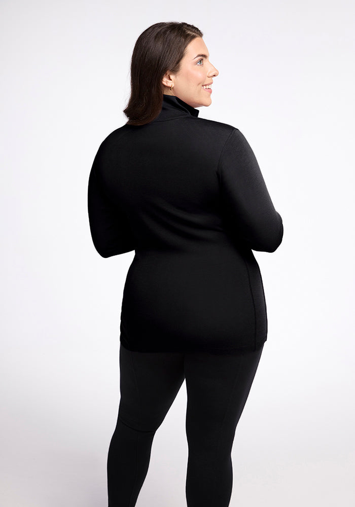 A woman with long dark hair is standing against a light grey background, facing away from the camera. She is wearing a black Woolx Brooke 1/4 Zip made of Merino wool and black pants. She is turned slightly to the side, looking back and smiling, showcasing the moisture-wicking fabric of her top.