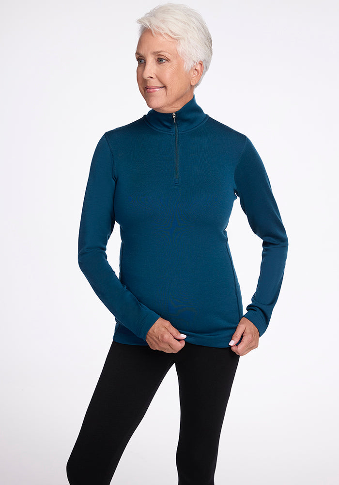 A woman with short white hair, dressed in Woolx's Brooke 1/4 Zip teal long-sleeved top and black pants, stands against a plain white background. She has a neutral expression and is holding the hem of her top with both hands.