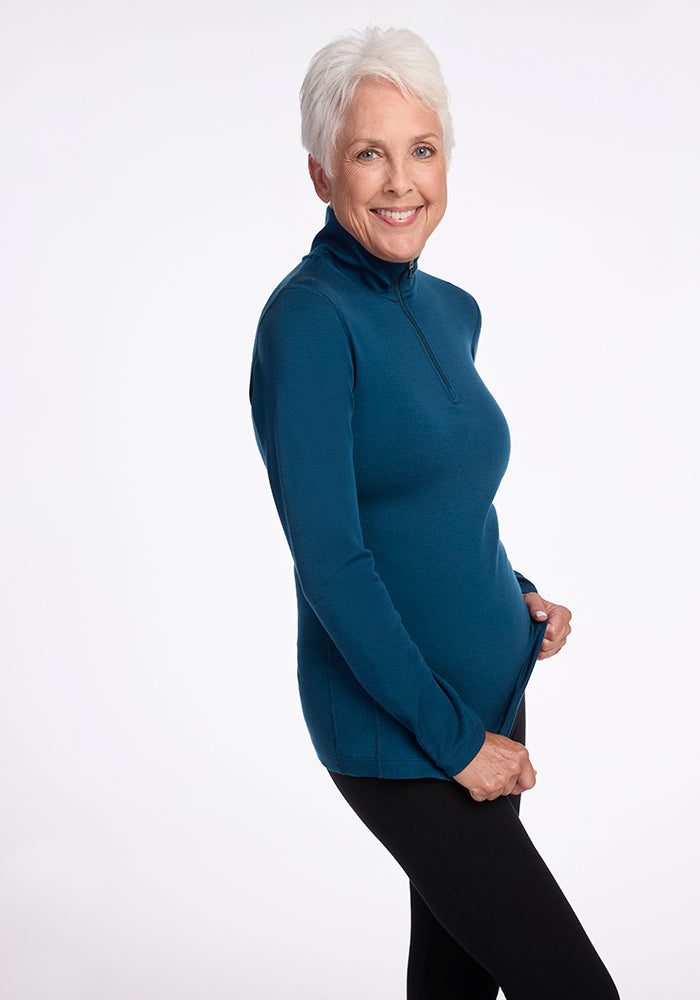 A smiling older woman with short white hair is dressed in the Woolx Brooke 1/4 Zip pullover and black leggings. She stands against a white background, holding the hem of her pullover with her left hand.