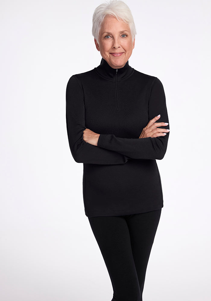 An older adult with short white hair is wearing the Woolx Brooke 1/4 Zip top and black pants, standing against a plain white background with arms crossed, smiling gently at the camera.