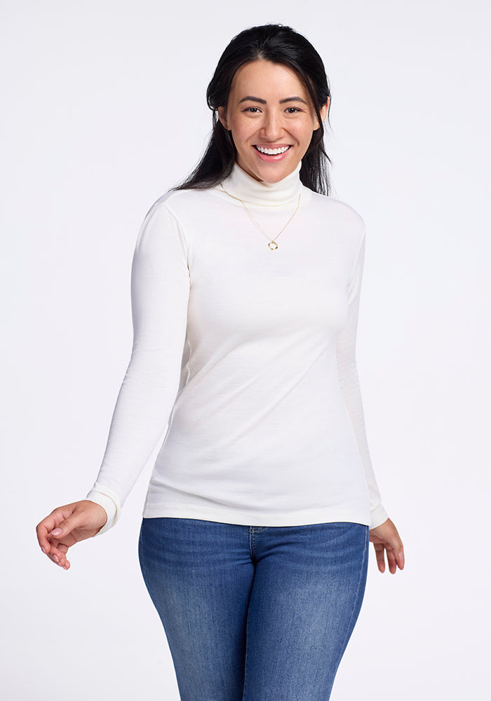 In a simple setting, a person smiles warmly in Woolx's Peyton Turtleneck in buttercream and blue jeans. Their long dark hair cascades over a delicate necklace, standing out against the plain white background.