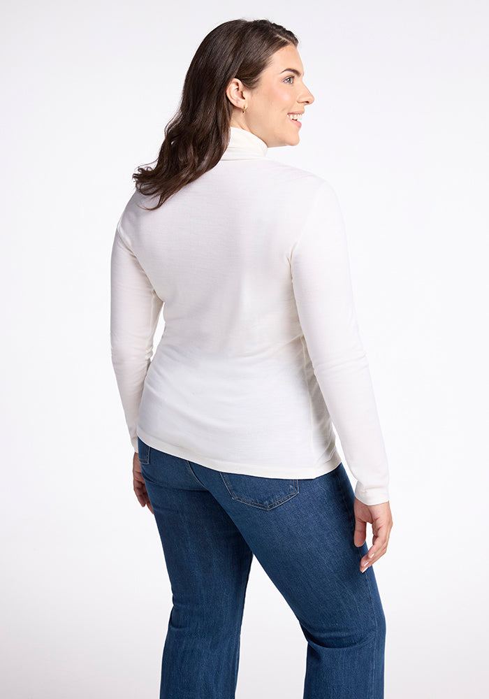 A woman with long brown hair is seen from the back, wearing a Woolx Peyton Turtleneck in buttercream and blue jeans. She is standing against a plain white background, ready for her next adventure.