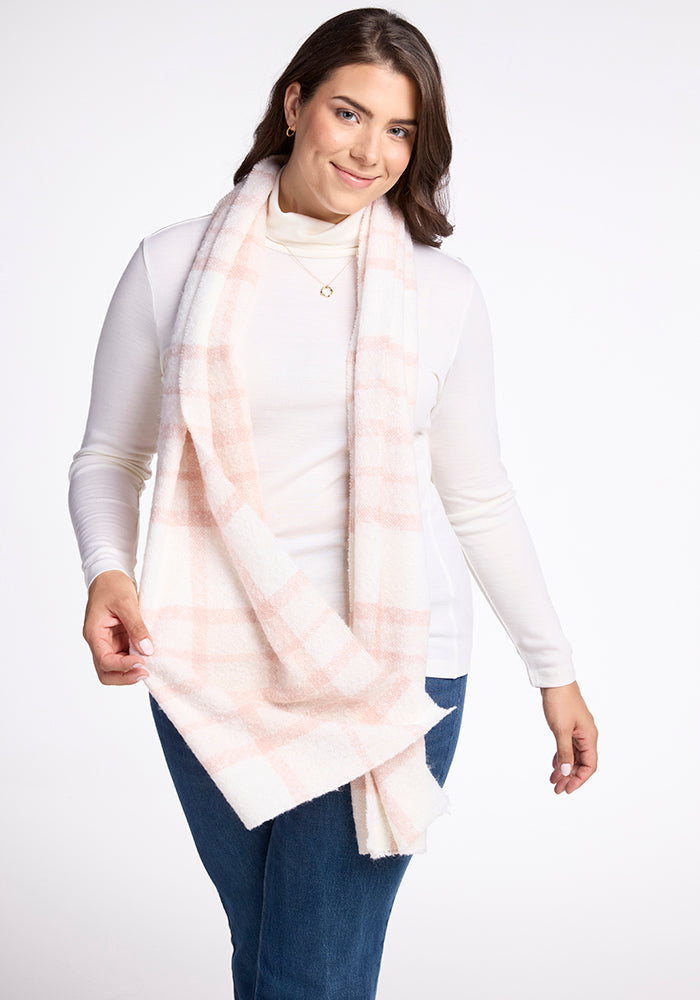 A person with shoulder-length dark hair is posing against a white background. They are wearing a long, mauve plaid scarf named the Bradie Scarf by Woolx, a white turtleneck, and blue jeans. They have a gentle smile and are holding the scarf with one hand.