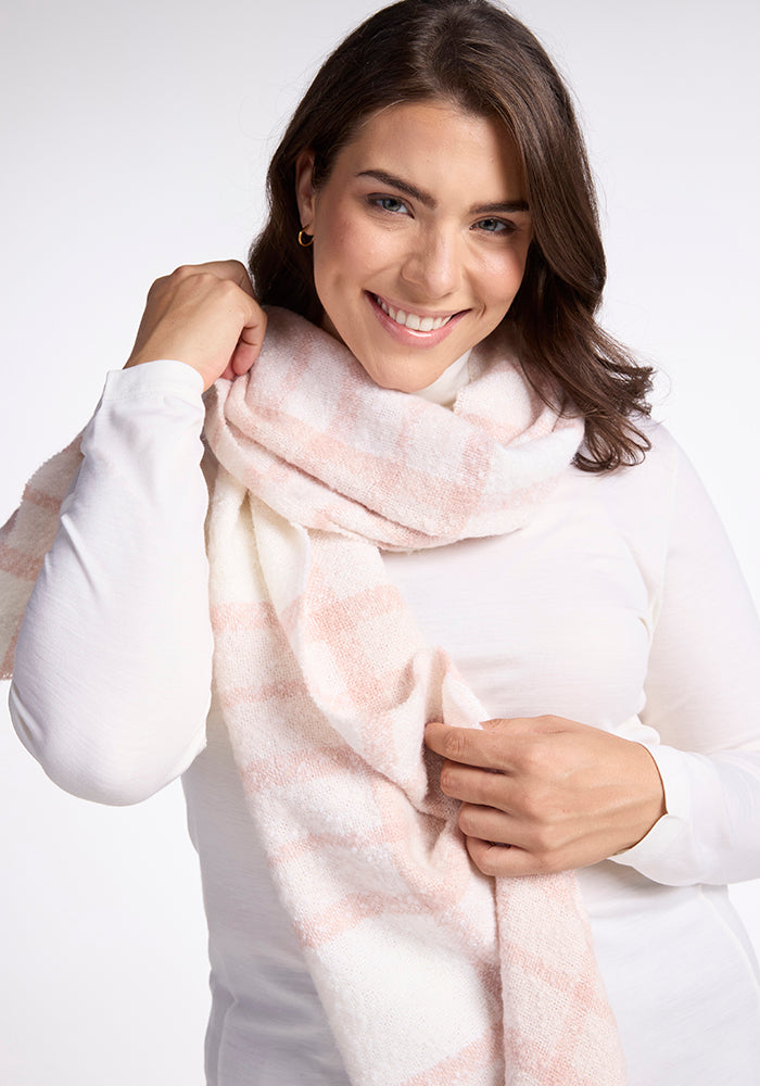 A person with shoulder-length dark hair smiles while wearing a white long-sleeve shirt and the Woolx Bradie Scarf in Mauve Plaid. They are adjusting the scarf with one hand and appear in front of a plain white background.