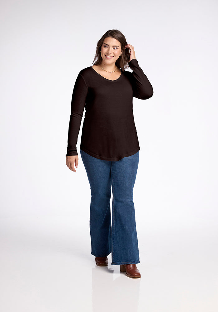 A woman with long brown hair is smiling and walking forward. She is wearing the Woolx Eva Tunic in French Roast, blue flared jeans, and brown ankle boots. With one hand near her hair, she embodies effortless street style against a plain white background.