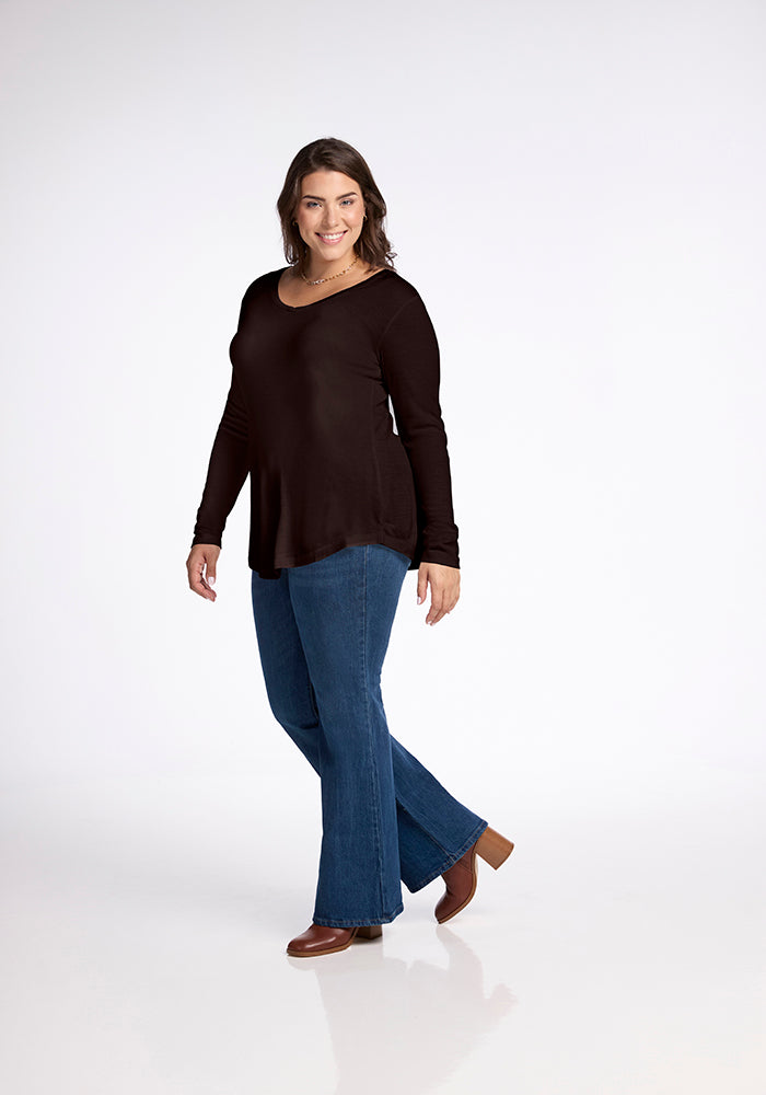 A woman with shoulder-length brown hair is smiling and walking towards the camera. She is wearing a Woolx Eva Tunic in French Roast, blue flared jeans, and brown heeled shoes. The background is white and uncluttered, highlighting her chic street style.