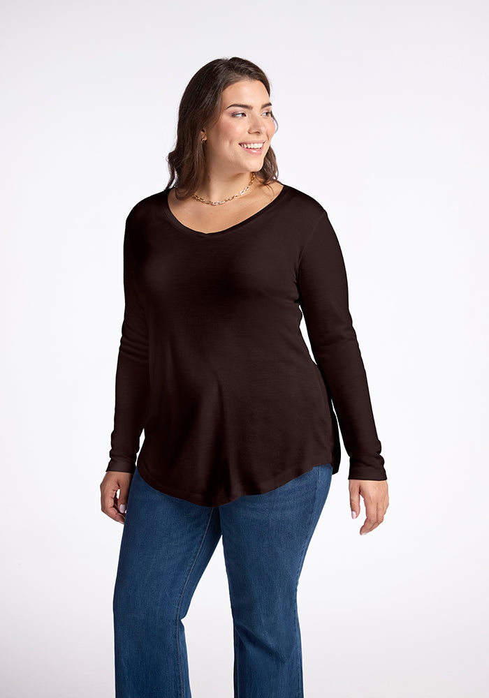 A woman with long dark hair smiles while looking to the side. She is wearing a French Roast Eva Tunic by Woolx and blue jeans, embodying effortless street style. The background is a plain white studio setting. 