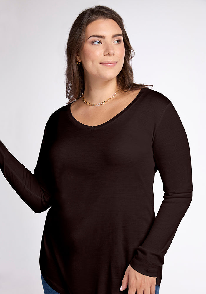 A woman with long brown hair is wearing the Eva Tunic in French Roast by Woolx and a gold necklace. She is standing against a plain, light-colored background, looking slightly to her right with a neutral expression.