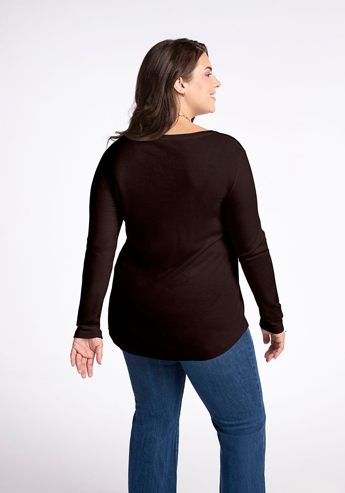 A woman with long dark hair, facing away from the camera and wearing a French Roast-colored Eva Tunic from Woolx along with blue jeans, stands against a white background. She is smiling slightly and showing an earring in her left ear.