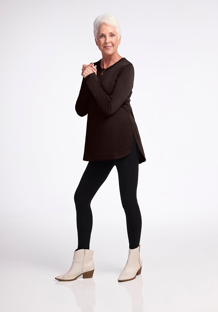 An older woman with short, white hair is standing against a plain background. She is wearing the Blair Tunic - French Roast from Woolx in a relaxed fit, paired with black leggings and light-colored ankle boots. Her hands are clasped together, and she is smiling at the camera.