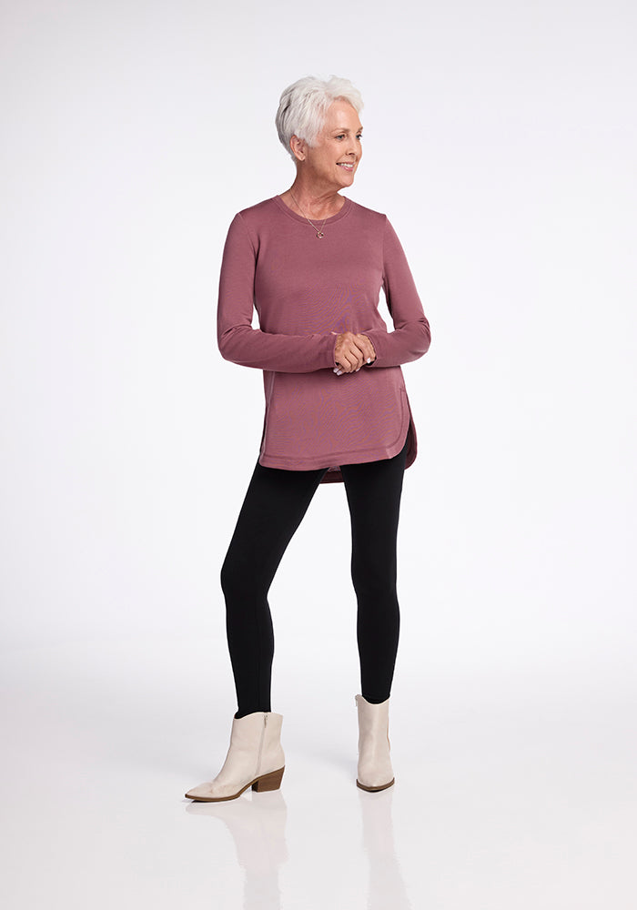 A woman with short white hair is standing against a plain background. She is wearing an ultra-cozy Woolx Blair Tunic in Wild Ginger made from Woolmark® Certified Australian Merino Wool, black leggings, and white ankle boots. Smiling and looking off to the side, she clasps her hands in front of her.