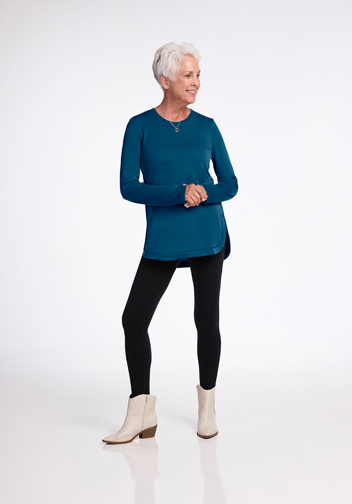 A person with short white hair is standing against a plain white background, wearing the ultra-cozy Woolx Blair Tunic in Real Teal, black leggings, and white ankle-length boots. They are smiling and have their hands loosely clasped in front of them.