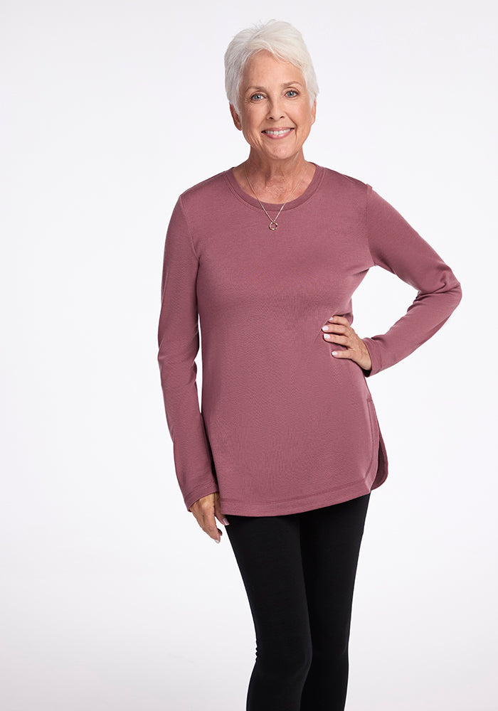 An elderly woman with short white hair stands against a plain background. She is wearing a long-sleeved, ultra-cozy Blair Tunic in Wild Ginger from Woolx and black pants. She smiles at the camera with one hand on her hip. 