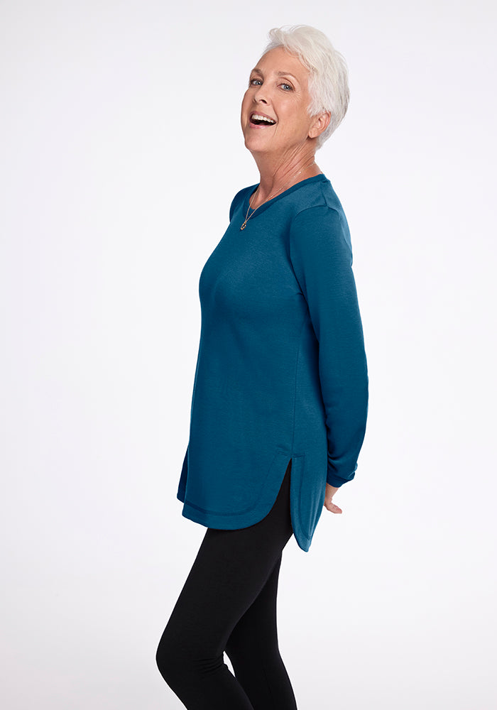 An older adult with short white hair is standing and smiling with their body turned slightly to the side. They are wearing an ultra-cozy Blair Tunic in Real Teal from Woolx and black leggings. The background is white. 