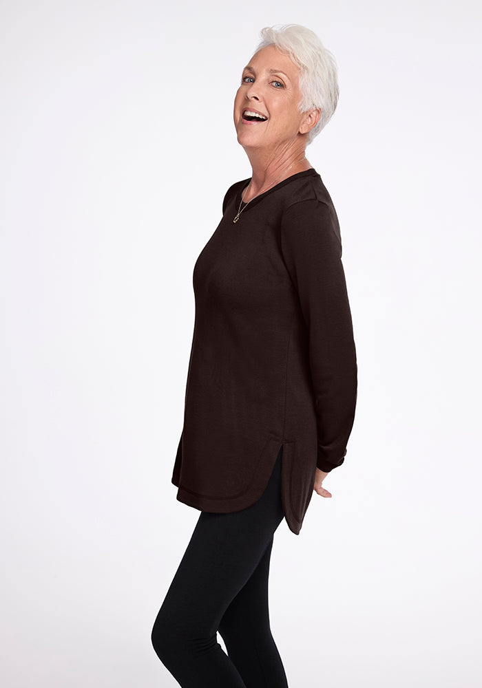 A smiling older adult with short white hair is standing and posing sideways, wearing a Woolx Blair Tunic in French Roast and dark pants. Their hands are clasped behind their back, and they have a cheerful expression on a plain, light background.