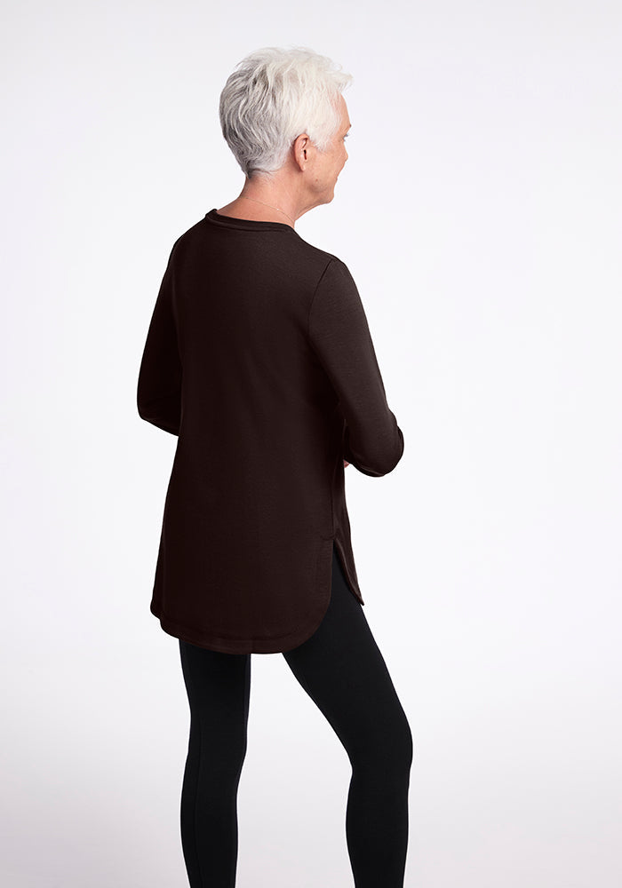 A person with short gray hair stands facing away from the camera, wearing a relaxed fit, long-sleeve Blair Tunic in French Roast by Woolx and black leggings. The background is plain white.