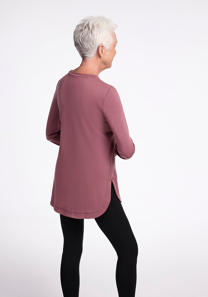 A person with short gray hair is standing against a plain background, facing away to the left. They are wearing an ultra-cozy, wild ginger Blair Tunic from Woolx, paired with black leggings. The relaxed fit of the tunic complements their style as they stand with arms crossed, looking to the side.