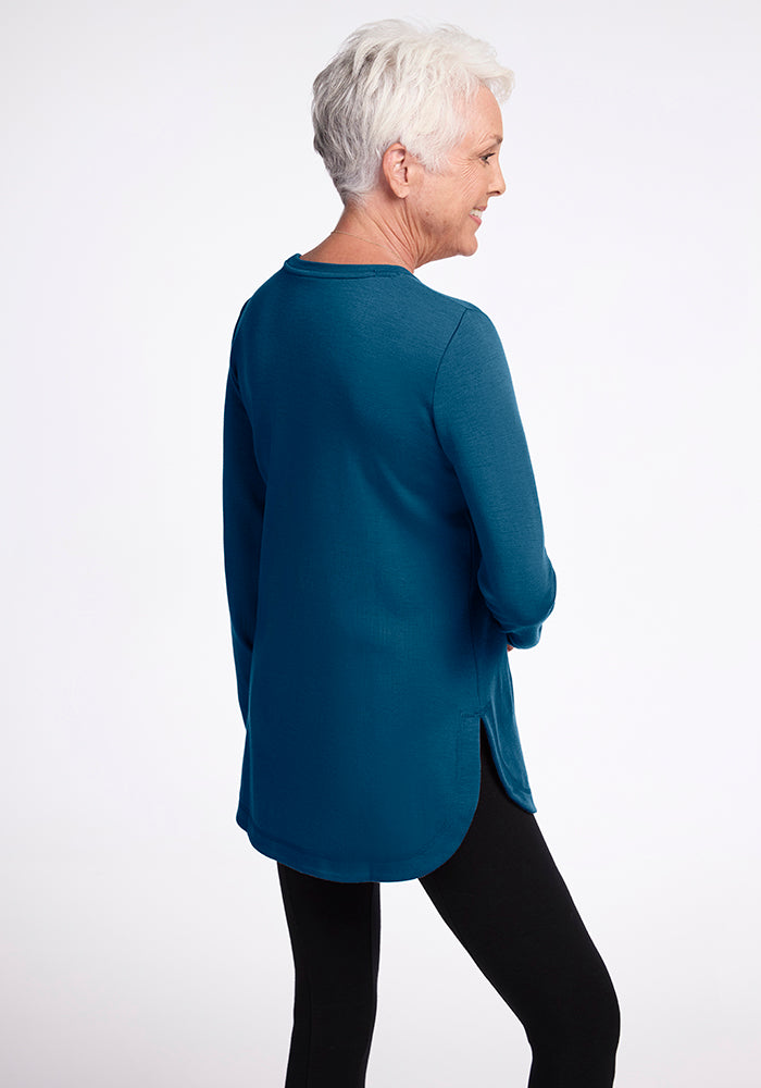 A woman with short white hair stands facing away from the camera, wearing a Woolx Blair Tunic in Real Teal, made from Woolmark® Certified Australian Merino wool and black leggings. Her relaxed posture and bent arm add to the simplicity of the plain white background.