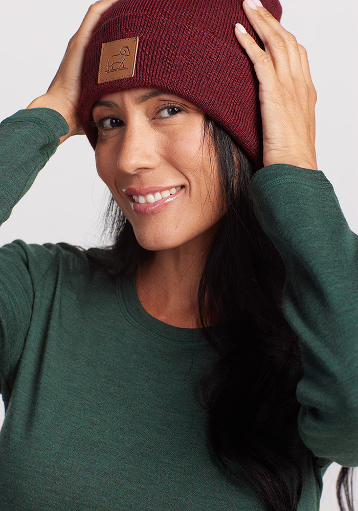 A person with long dark hair wearing a green long-sleeve shirt and a Woolx Baylor Beanie in Cranberry Melange is smiling and holding the sides of the beanie. The Merino wool beanie features a small rectangular patch with a simple line drawing of a bird. The background is plain and light-colored.