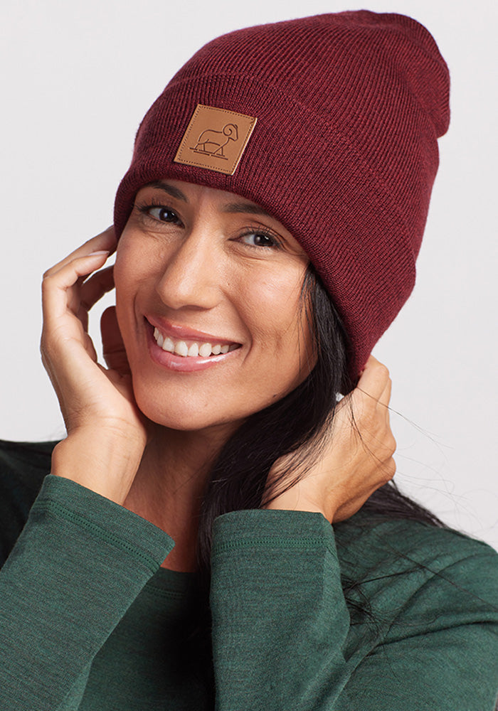 A person with long dark hair smiles at the camera while wearing a Woolx Baylor Beanie in Cranberry Melange, adorned with a tan patch featuring a simple animal outline. Dressed in a green top, they have their hands near their face and pose cheerfully, showcasing their stylish and warm hat.