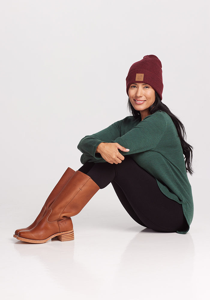 A person with long dark hair, wearing a cranberry melange Baylor Beanie from Woolx, a green Merino wool sweater, black leggings, and brown knee-high boots sits on the floor. Their legs are bent, arms resting on their knees, and they are smiling against a plain white background.