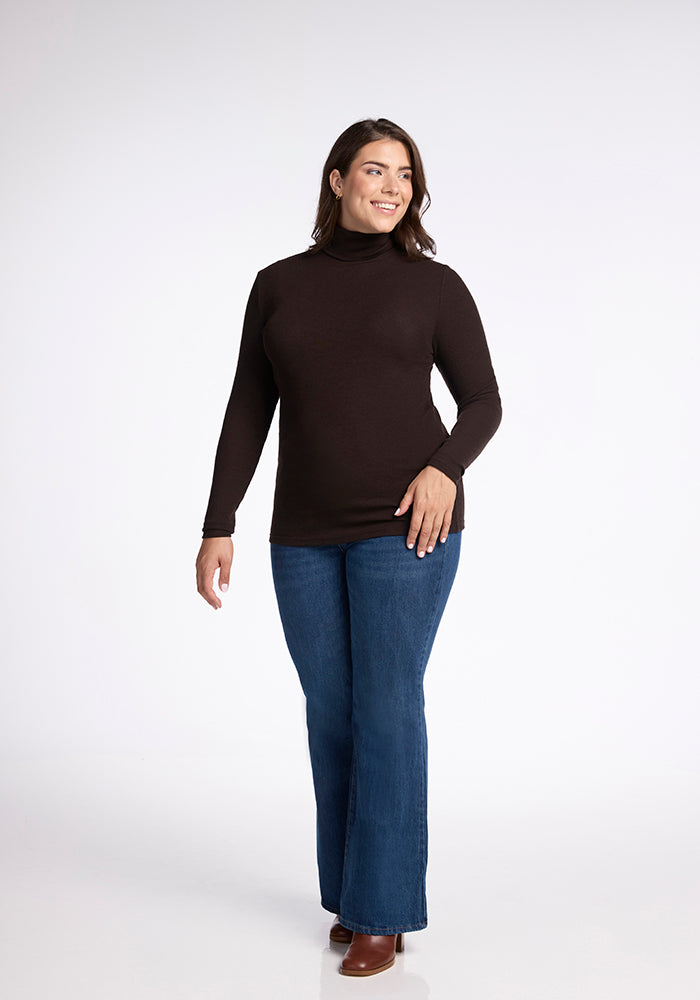 A woman with long hair smiles while wearing a moisture-wicking, long-sleeved Sage Turtleneck by Woolx, blue jeans, and brown shoes. She stands against a plain white background.