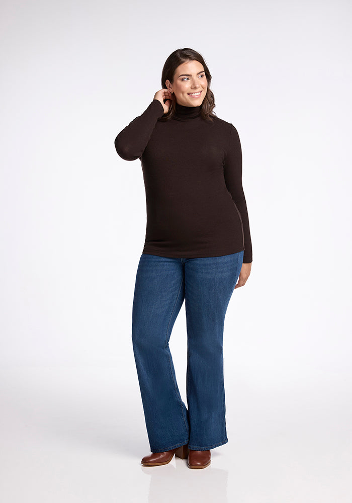 A woman with medium-length dark hair stands against a white background. She is wearing a long-sleeved Sage Turtleneck from Woolx, blue flared jeans, and brown shoes. She is smiling and has one hand raised to her ear, appearing relaxed and comfortable.