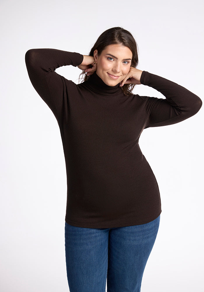 A person wearing a Sage Turtleneck by Woolx and blue jeans poses with their hands behind their head, smiling confidently against a plain white background. 