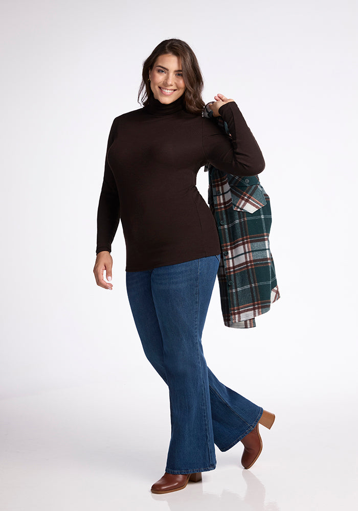 A woman is standing in a studio, smiling. She has long dark hair and is wearing a fitted black Sage Turtleneck from Woolx, blue bell-bottom jeans, and brown shoes. She holds a green and white plaid coat over her shoulder with one hand. The background is white and uncluttered.