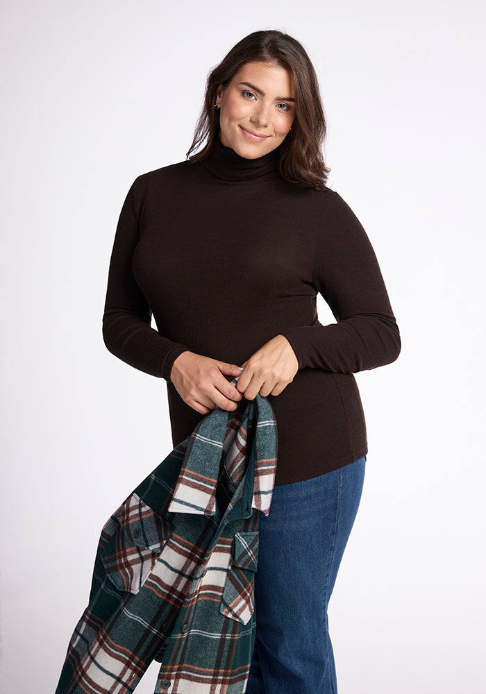 A person with shoulder-length hair is wearing a long-sleeved Sage Turtleneck from Woolx and blue jeans. They are holding a green and white plaid shirt in both hands, standing against a plain white background.