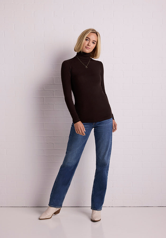 A person with short blonde hair stands against a white brick wall, wearing a dark long-sleeve Sage Turtleneck from Woolx, blue jeans, and white heeled boots. They have a relaxed posture, with one leg slightly forward and one hand resting on their hip, smiling gently.