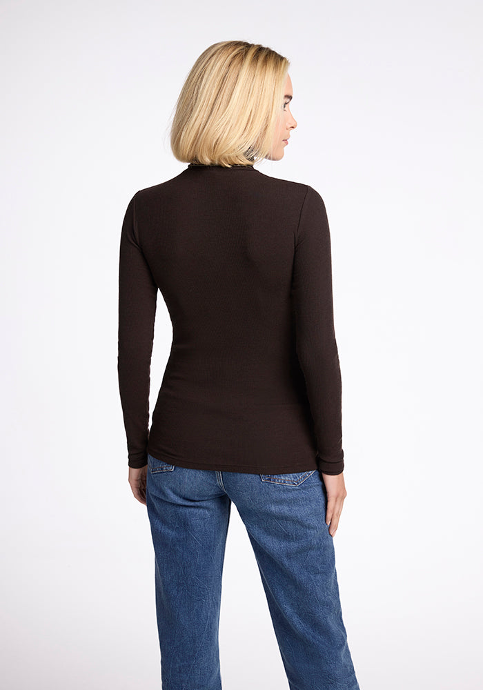 A person with short blonde hair is standing with their back facing the camera. They are wearing a long-sleeved, dark-brown Sage Turtleneck from Woolx and blue jeans, ensuring long-lasting comfort. The background is plain white.