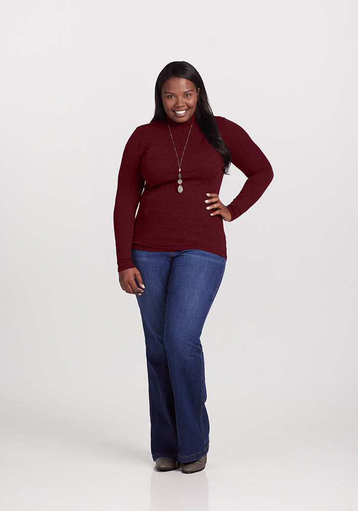 A woman is standing against a plain white background, wearing a long-sleeved maroon Sage Turtleneck from Woolx, praised for its long-lasting comfort, blue jeans, and brown shoes. She has a silver necklace and a smile on her face, with her hands resting on her hips.