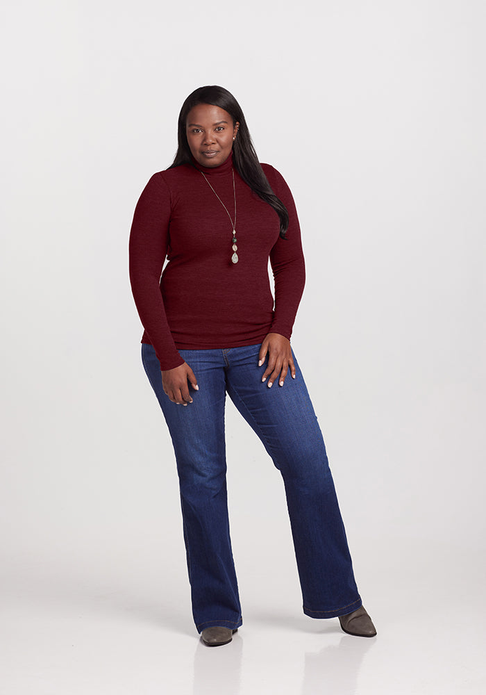 A woman is standing against a plain background, wearing a maroon Sage Turtleneck from Woolx, blue jeans, and grey shoes. She has long, dark hair and is accessorized with a long necklace. She is posed with one hand in her pocket and has a confident expression.