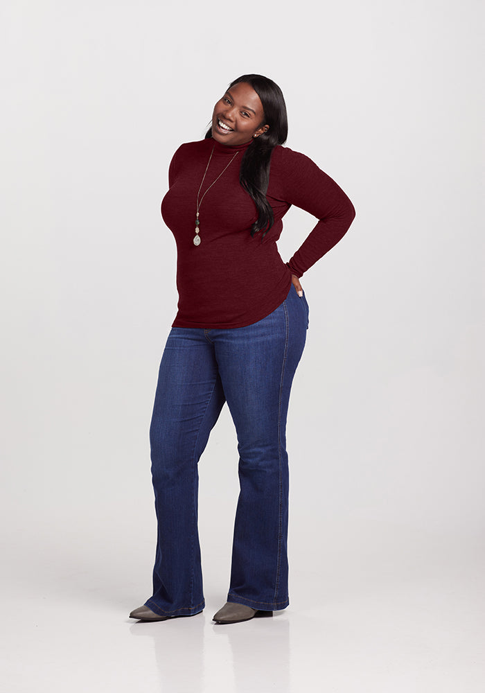 A woman smiles and poses confidently. She is wearing a long-sleeved, dark red Sage Turtleneck by Woolx, blue jeans, and gray shoes. She accessorizes with a long necklace featuring pendant details. Her hands rest on her hips as she stands against a plain white background.