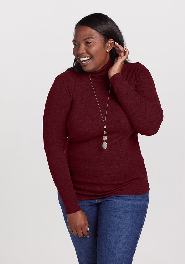 A person with long dark hair smiles and looks to the side, wearing a maroon Sage Turtleneck from Woolx and blue jeans. They have a long pendant necklace and are lifting one hand near their ear. The background is plain white. 