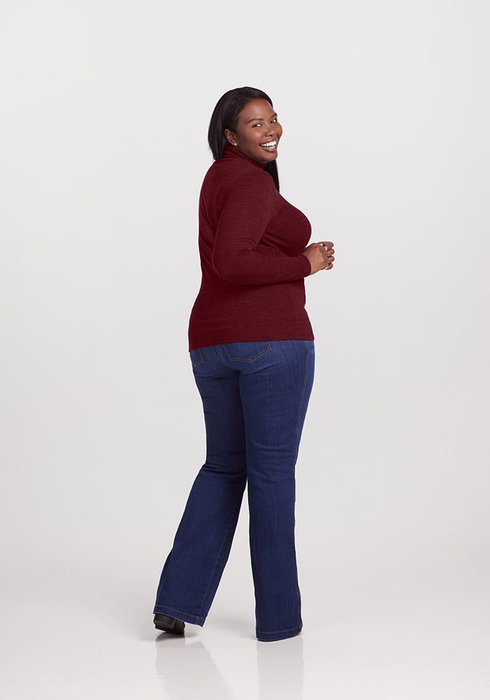 A woman stands smiling and looking over her shoulder. She is wearing a maroon Woolx Sage Turtleneck, blue jeans, and black shoes. The background is plain and light gray.